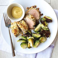 a white plate topped with meat and brussel sprouts next to a bowl of mustard