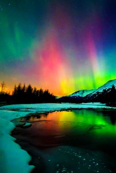 an aurora bore is seen over the water in this colorful sky with snow and trees