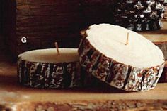 two candles sitting next to each other on top of a wooden table with pine cones in the background