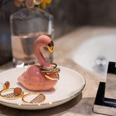 a pink flamingo on a white plate next to a glass of water and gold rings