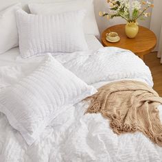 a bed with white comforters and pillows on top of it next to a table