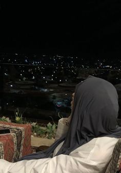 a woman sitting on top of a chair next to a window at night with the city lights in the background