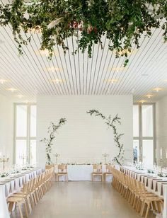 an indoor wedding venue with white walls and greenery hanging from the ceiling, long tables set for guests