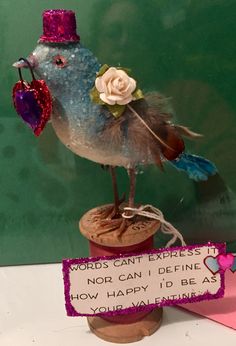 a blue bird with a pink hat and heart on it's head sitting on top of a wooden post