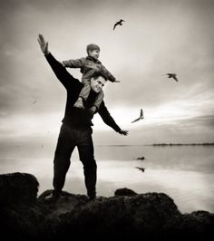 a black and white photo of two people standing on rocks with birds flying in the background