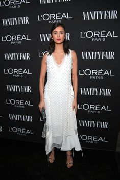 a woman standing in front of a black wall wearing a white dress and holding a purse