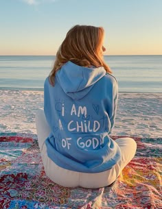 a woman sitting on the beach wearing a hoodie that says i am a child of god