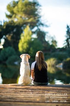 a woman sitting next to her dog on the dock looking out at the water with trees in the background