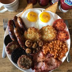 an english breakfast with eggs, sausages, beans and bacon on a plate next to coffee