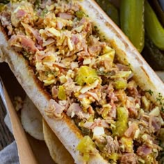 a hot dog with toppings sitting on top of a wooden tray next to pickles