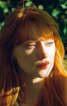 a young woman with red hair and freckled bangs looks into the camera while standing in front of trees