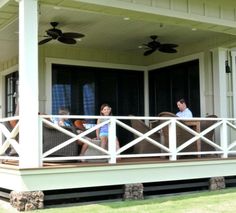 three people sitting on the porch of a house with ceiling fans in the back ground