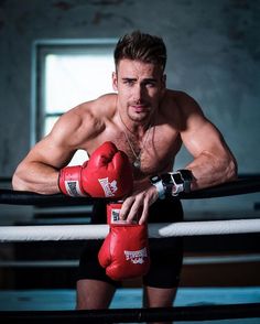 a shirtless man leaning on a boxing ring with his hands on the ropes and looking at the camera