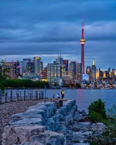 the city skyline is lit up in red, white and blue as people sit on rocks near the water