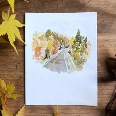 a watercolor painting of a road surrounded by autumn leaves on a wooden table with fall foliage