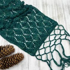 a pine cone is next to a green knitted scarf on a white wooden surface