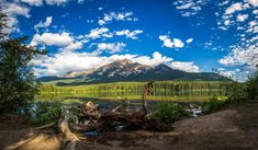 a lake surrounded by trees and mountains under a blue sky with puffy white clouds