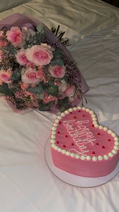 a pink heart shaped cake next to a bouquet of flowers on a white tablecloth