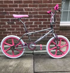 a pink and silver bicycle parked in front of a red brick wall with white spokes