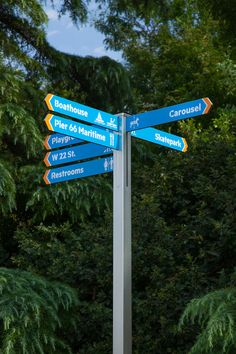 street signs pointing in different directions on a pole near some trees and bushes at an intersection