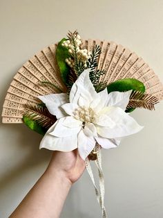 a hand holding a white flower on top of a green leafed fan with greenery