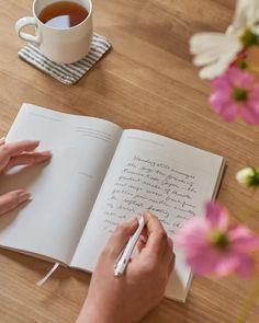 a person writing on a book with a cup of tea and flowers in the background