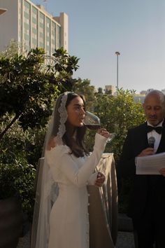 a woman in a wedding dress drinking from a wine glass while standing next to an older man