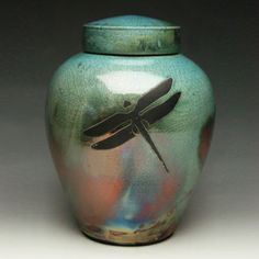 a green vase with a dragonfly painted on it's side and lid sitting in front of a white background