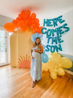 a pregnant woman standing in front of balloons and a sign that says here comes the son