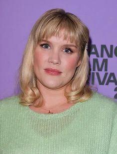 a woman with blonde hair wearing a green sweater and looking at the camera while standing in front of a purple wall