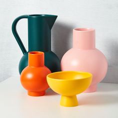 three different colored vases sitting next to each other on a white counter top with a gray wall in the background