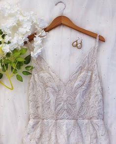 a white dress hanging on a wooden hanger next to some flowers and a pair of earrings