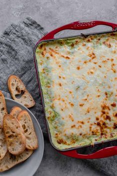 a red casserole dish with cheese and bread on the side