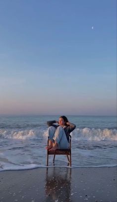 a woman sitting in a chair on top of a beach next to the ocean under a blue sky