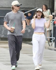 a man and woman walking down the street while talking on their cell phones, holding drinks