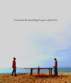 two men are standing on the beach near a picnic table with an ocean in the background