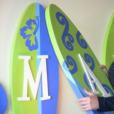 a woman holding two surfboards in front of a wall with the letter m on it