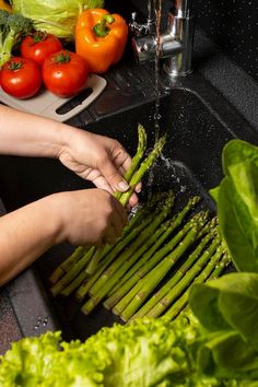 a person washing asparagus in a sink with tomatoes, lettuce and other vegetables