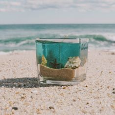 an empty glass filled with sand and sea shells on top of a sandy beach next to the ocean