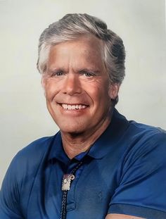 an older man with grey hair and blue shirt smiling at the camera while wearing a tie