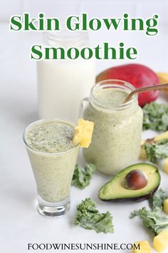 two glasses filled with green smoothie next to an avocado and other fruits