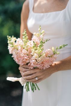 a woman in a white dress holding a bouquet of pink carnations and greenery