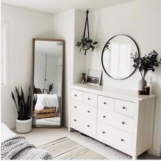 a bedroom with white furniture and plants on the dresser next to a large round mirror