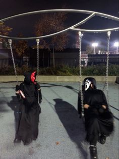 two people dressed in black sit on swings at night with their faces painted red and white