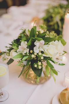 white flowers and greenery in a glass vase on a table with candles, plates and napkins