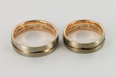 two gold wedding rings with engraved names on the sides and hearts in each ring, sitting side by side