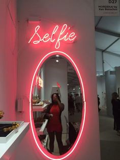 a woman taking a selfie in front of a pink neon sign that says selfie