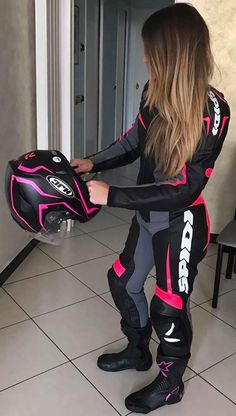 a woman in black and pink riding suit holding onto her helmet while standing on tiled floor