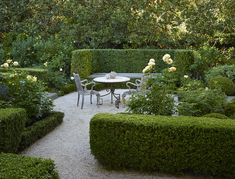 a table and chairs are in the middle of a garden with hedges on both sides