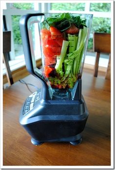 a blender filled with lots of vegetables on top of a wooden table next to a window